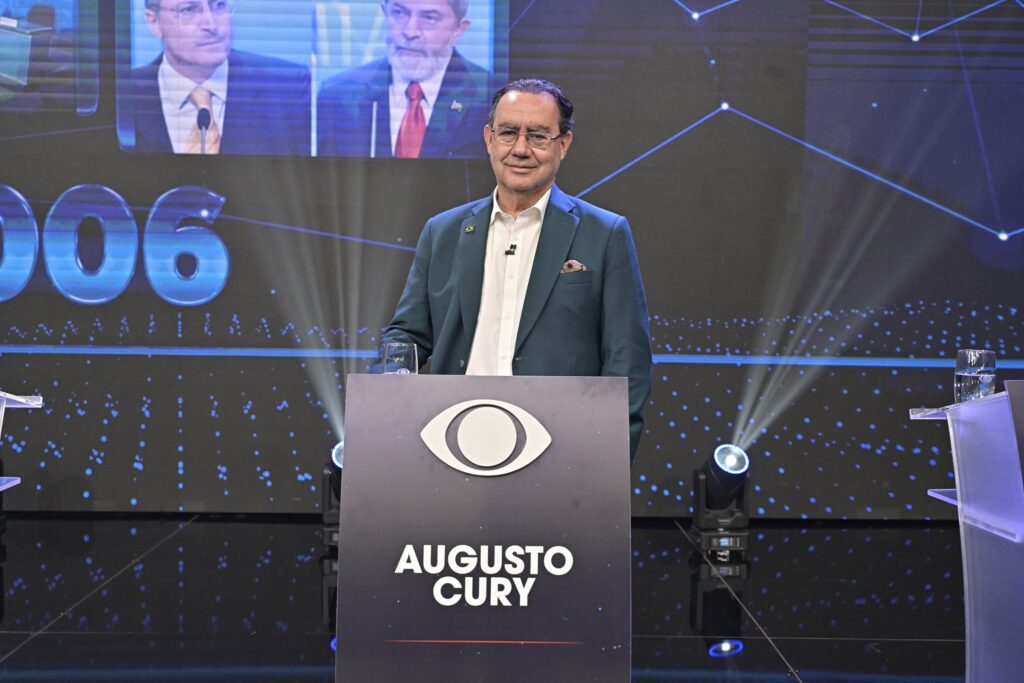 Augusto Cury está de pé atrás de um púlpito escuro com o logotipo da Band e o nome "Augusto Cury" impresso. Ele veste um paletó azul-petróleo sobre camisa branca sem gravata e usa óculos de grau. Ao fundo, há um telão com iluminação azul e um painel com imagens de outros candidatos.