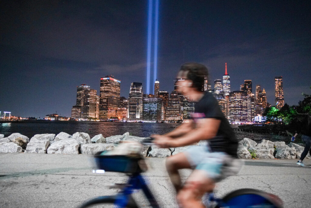 Menino caminha de bicicleta. Ao fundo, luzes iluminam local onde estavam as Torres Gêmeas, em Nova York | Foto: REUTERS/Ryan Murphy